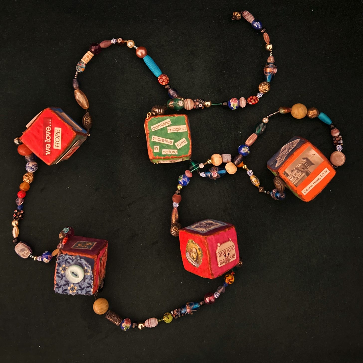 Five papier-mâché cubes joined together on beaded strings. The cubes are highly coloured and have images and text stuck to them.