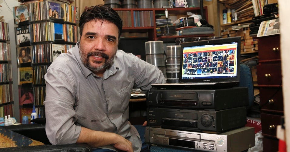 homem de barba e cabelos negros em uma sala cheia de cds e dvds. ele está ao lado de aparelhos vhs empilhados, e sobre esta pilha, um notebook aberto exibe uma página com pequenas imagens.