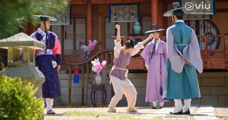 A woman in peasant clothes dances triumphantly amid three men in fancy hanbok