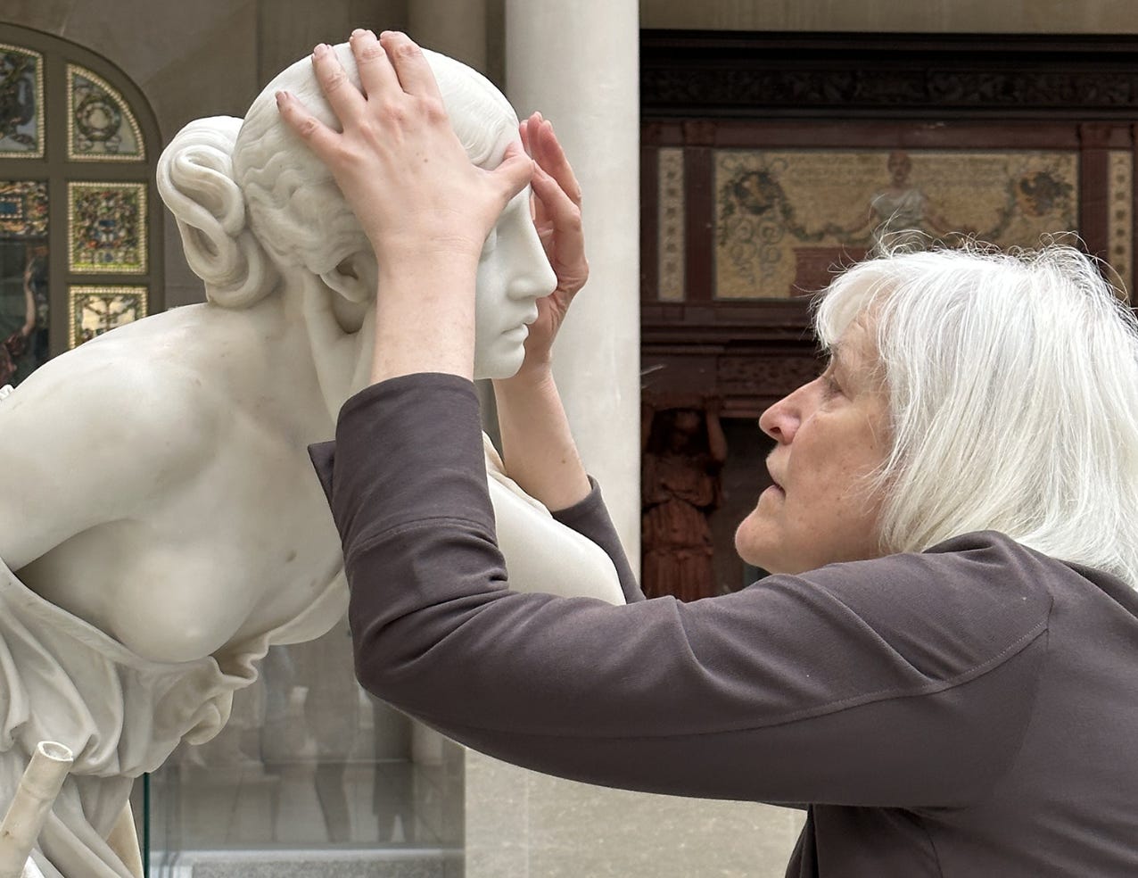 Image of the writer and professor Georgina Kleege, with her hands around the head of Nydia, The Blind Flower Girl of Pompeii, during a touch tour in the American Wing Engelhard Sculpture Court at The Met, a skylit space with direct, dramatic natural light. The image depicts the profile of a grey-white-haired woman with medium long straight hair with a long-sleeved brownish purple sweater touching the face of an almost life-sized white marble statue of a young girl. Her left hand is on the top of the sculpture's head, and her right thumb is touching the forehead. Two decorative artworks are installed into the wall in the background: one stained glass window and a fireplace.