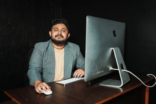 Free Smiling man in office attire working at a desk using a computer and wireless mouse. Stock Photo