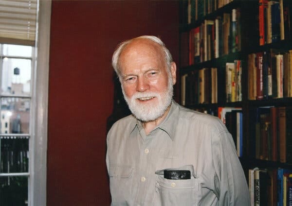 A casually dressed Mr. Fancher, with white hair and a full beard, standing in front of a bookcase and smiling.