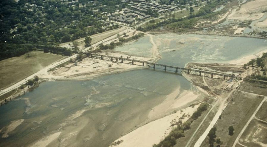 The image shows the Arkansas River at 29th Street and Riverside Drive in Tulsa, Oklahoma, during construction of the original Zink Dam. The river is wide and shallow, an earthy greyish green color, with sandbars drifting throughout the riverbed at varying depths and a wide exposed sandbar in the foreground on the west (right) bank of the river. The image is taken from north of the construction site and bridge, high above the west bank of the river. Triangular construction areas surrounded by earthen cofferdams occupy much of the riverbed beneath either end of the original pedestrian railroad bridge, which is located just upstream of the dam construction. Beyond the east (left) bank of the river are the River Parks trail; Riverside Drive; a small parking lot and large grassy field intersected by the Midland Valley trail (now the site of Gathering Place); the tree-lined streets of Maple Ridge; and a comparatively barren looking apartment complex just south of 31st street. Beyond the west (right) bank of the river is the river trail and a sparsely vegetated area, all of which is contaminated land held by the refinery. Some distance downstream of the construction site, an earthen barrier spans the riverbed in a wide arc, separating the impounded area below the bridge and dam site from a much drier, rockier riverbed below.