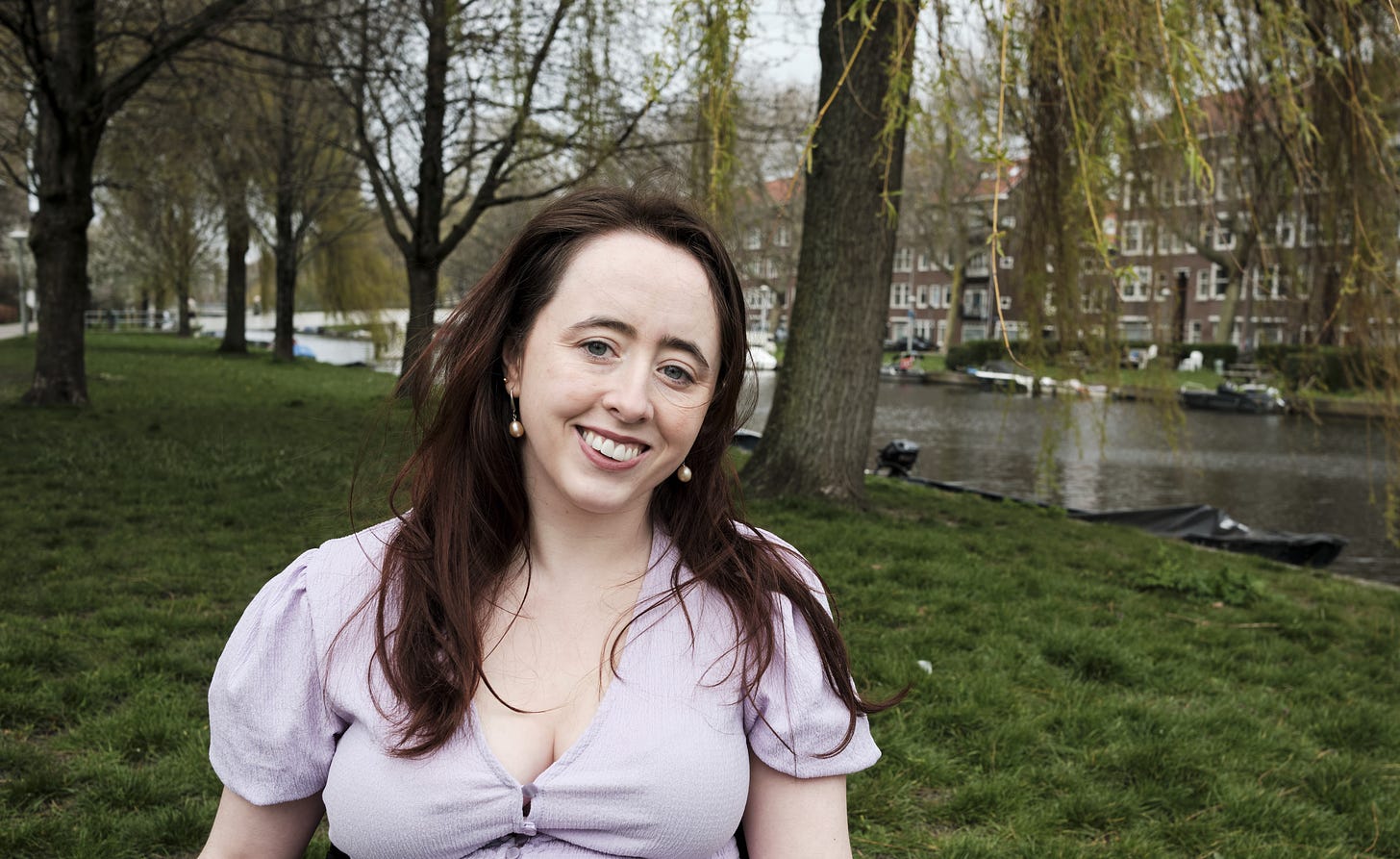 Niamh, an Irish woman with long dark brown hair is wearing a mauve top and smiling into the camera. She is sitting in a garden with trees lining a water body. Brown buildings with windows can be seen in the background. 
