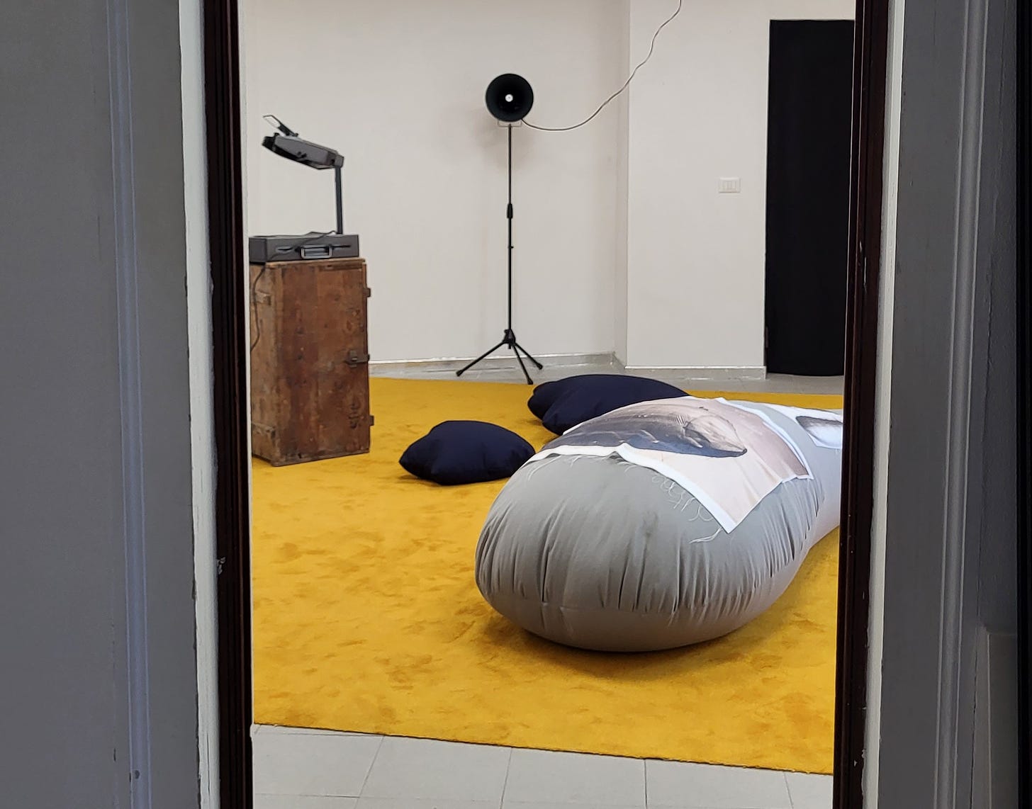 A white room with a yellow carpet, blue pillows, a grey fabric object, an overhead projector on a wooden box, and a speaker.