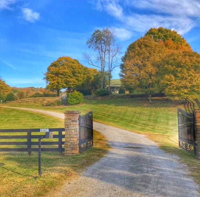 An open iron gate and a gravel driveway lead up to a farmhouse partially obscured by trees.