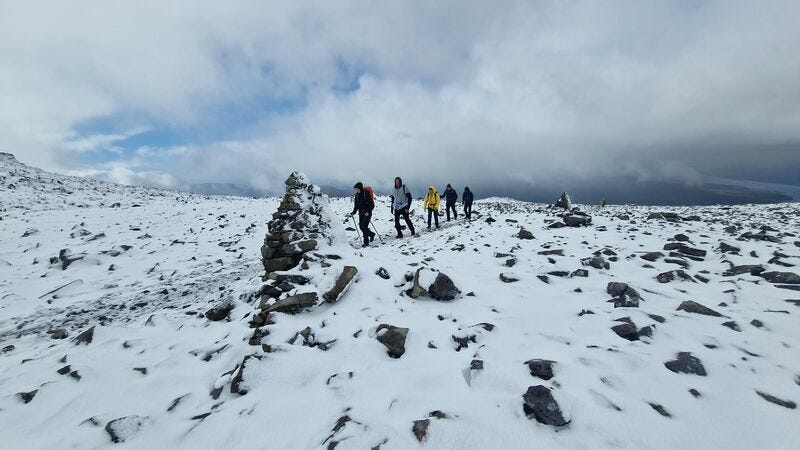 Five people walk across from right to left in the middle of the photo. They are about to pass behind a large cairn, with snow plastered on one side. The foreground is snowy with rocks sticking through and there are moody clouds in the background with a small patch of blue sky showing. Five people walk across from right to left in the middle of the photo. They are about to pass behind a large cairn, with snow plastered on one side. The foreground is snowy with rocks sticking through and there are moody clouds in the background with a small patch of blue sky showing.
