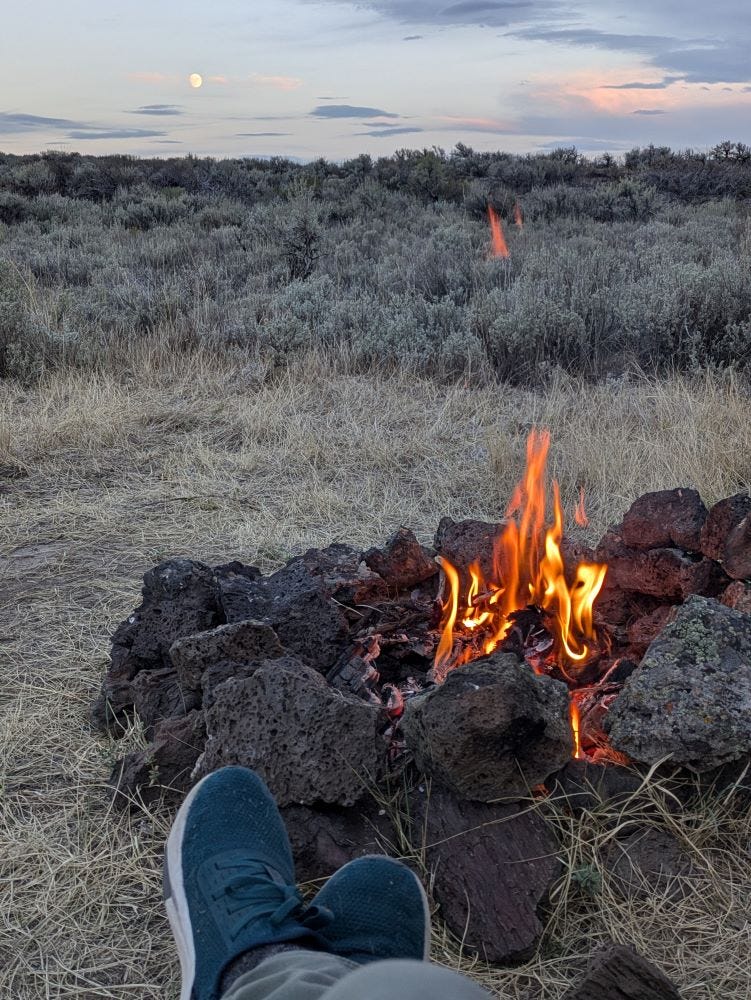 a view of my feet in tennis shoes, crossed in front of a small campfire with the desert sage and a twilight sky of pink and pale blue, an almost full moon just above the horizon