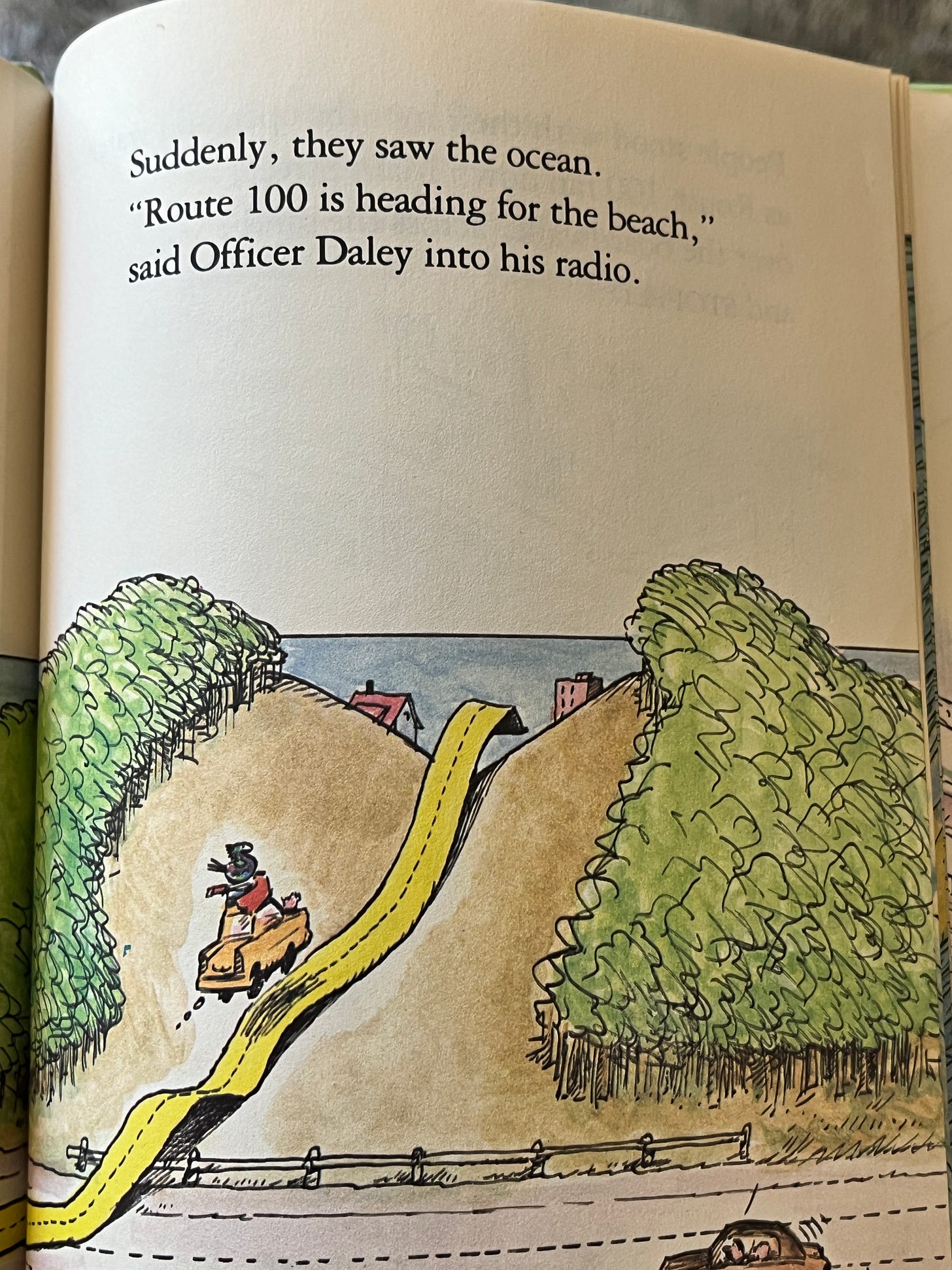 A page from the book. At the top it says "Suddenly, they saw the ocean. Route 100 is heading for the beach," said Office Daley into his readio." The bottom half of the page is an illustration of a steep hill with grass or trees on either side. A yellow road goes up and over the hill. Along the horizon is a plain blue ocean.