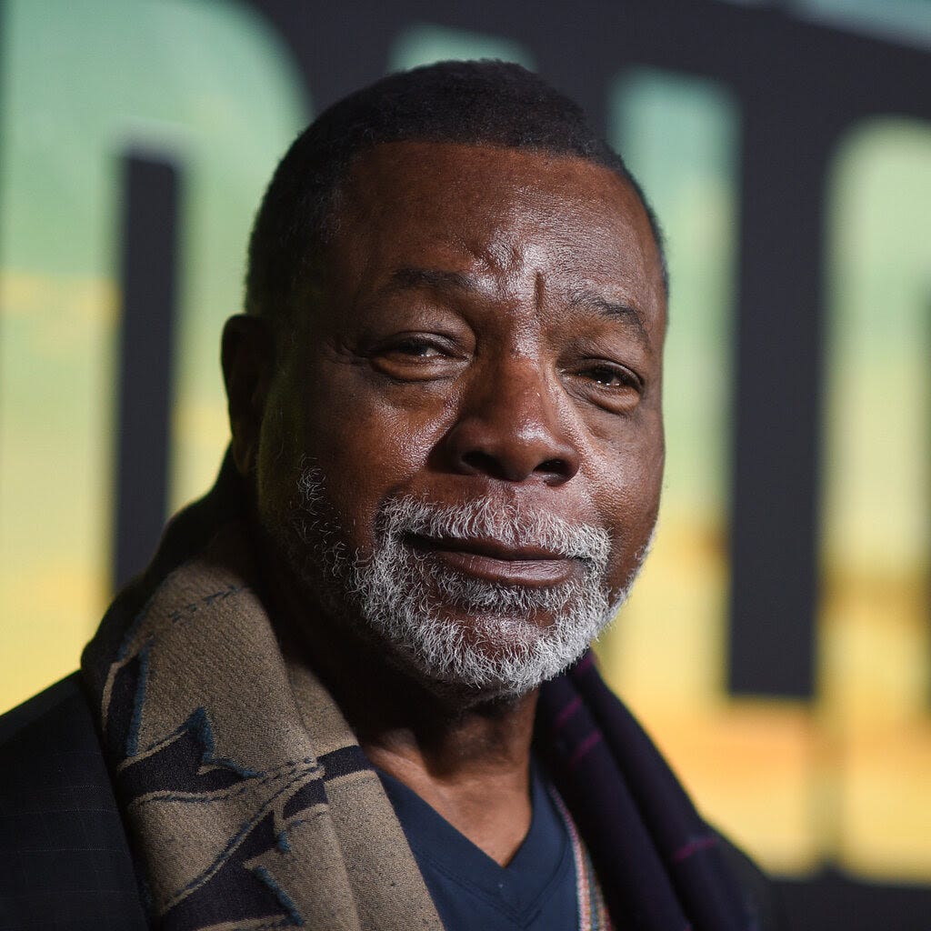 Carl Weathers looking at the camera. He has a gray beard and wears a tan scarf.