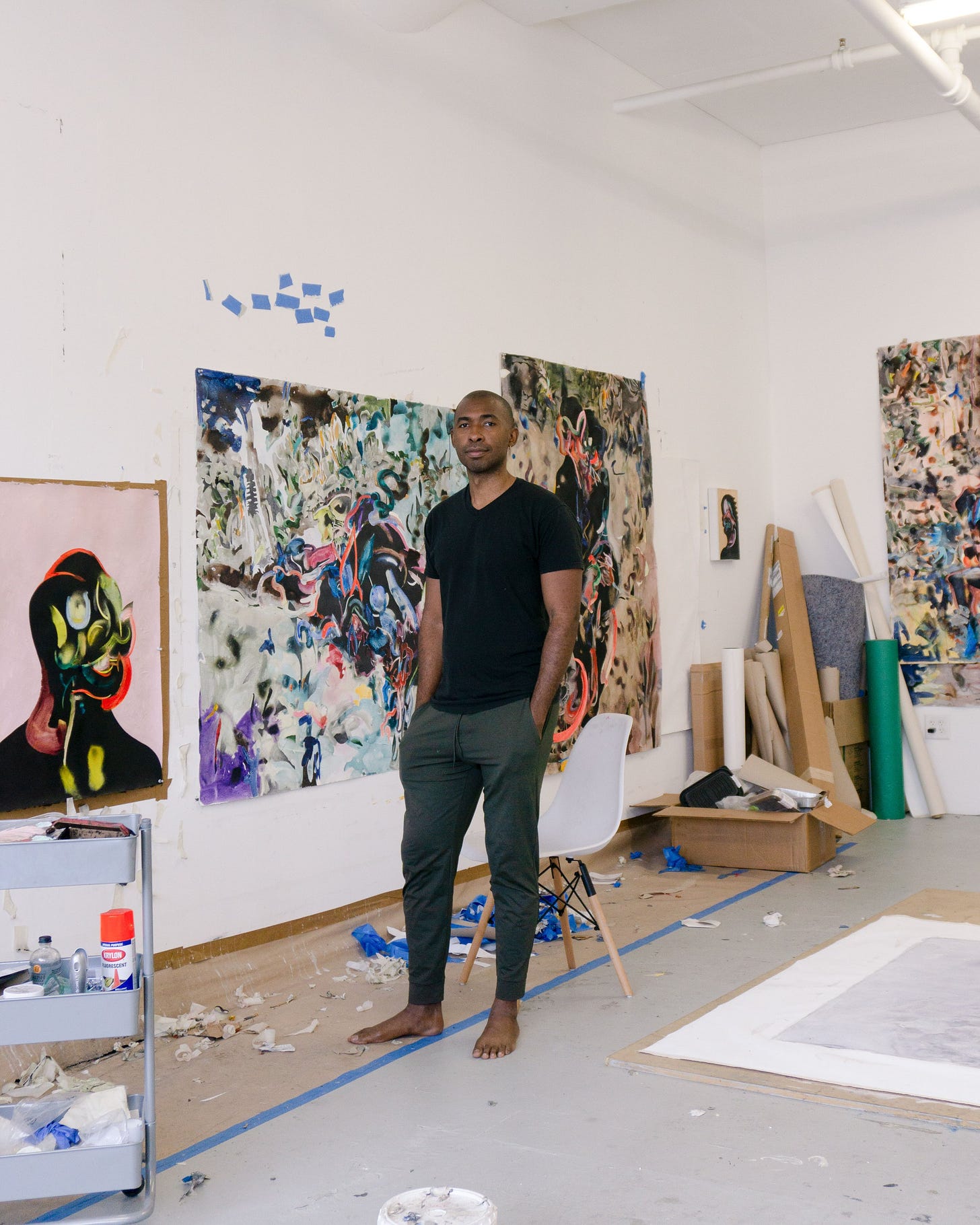 A Black person stands in a studio with his hands in his pockets. He is wearing a black T shirt and gray pants, and he is barefoot. To the right of him, colorful paintings are hung on the walls. The paintings are pink, blue, white, tan, and orange, a mix of abstract paint strokes and human like forms. 