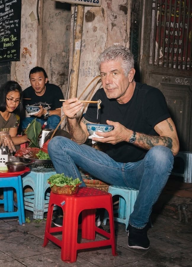 This may contain: a man eating food with chopsticks while sitting on stools in front of other people