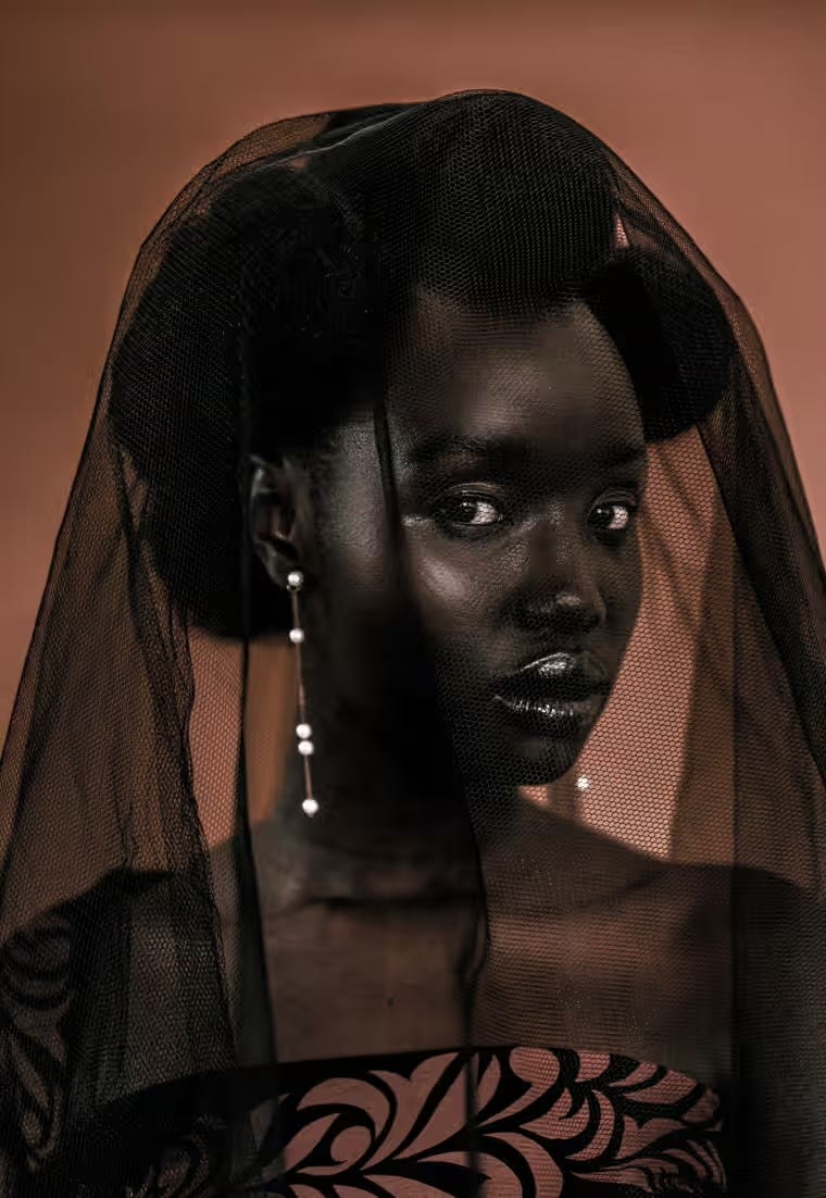 A sepia-toned portrait of an elaboratedly made-up African woman with her hair in dark coils, wearing a black lace veil