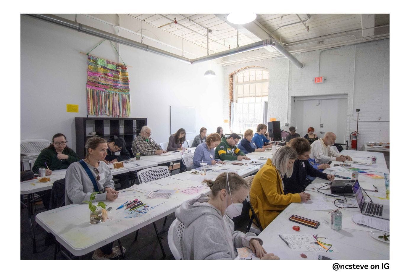 A full class picture. Everyone is working on their still life drawing warm-ups at long white tables with art supplies scattered around. There is a large window emitting light and a large colorful tapestry hanging on the wall behind the class.