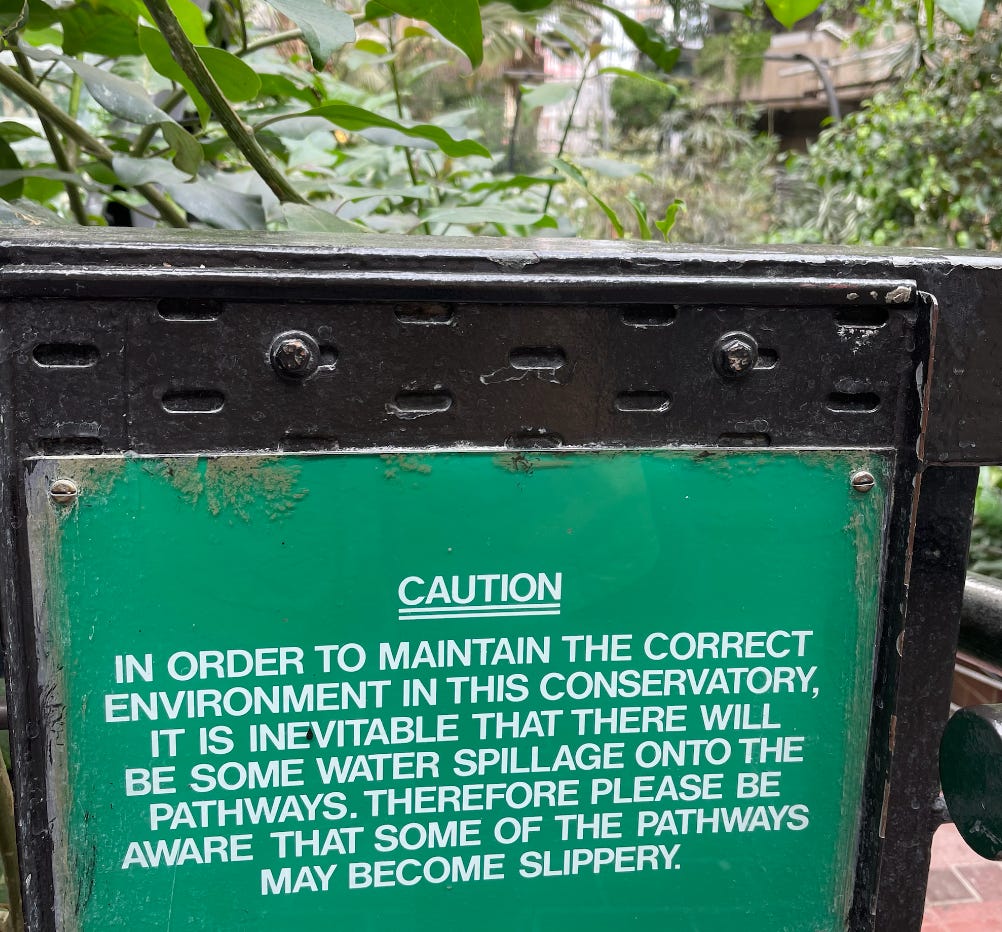 A green sign with white text that says: Caution: In order to maintain the correct environment in this conservatory, it is inevitable that there will be some water spillage onto the pathways. Therefor please be aware that some of the pathways may become slippery.