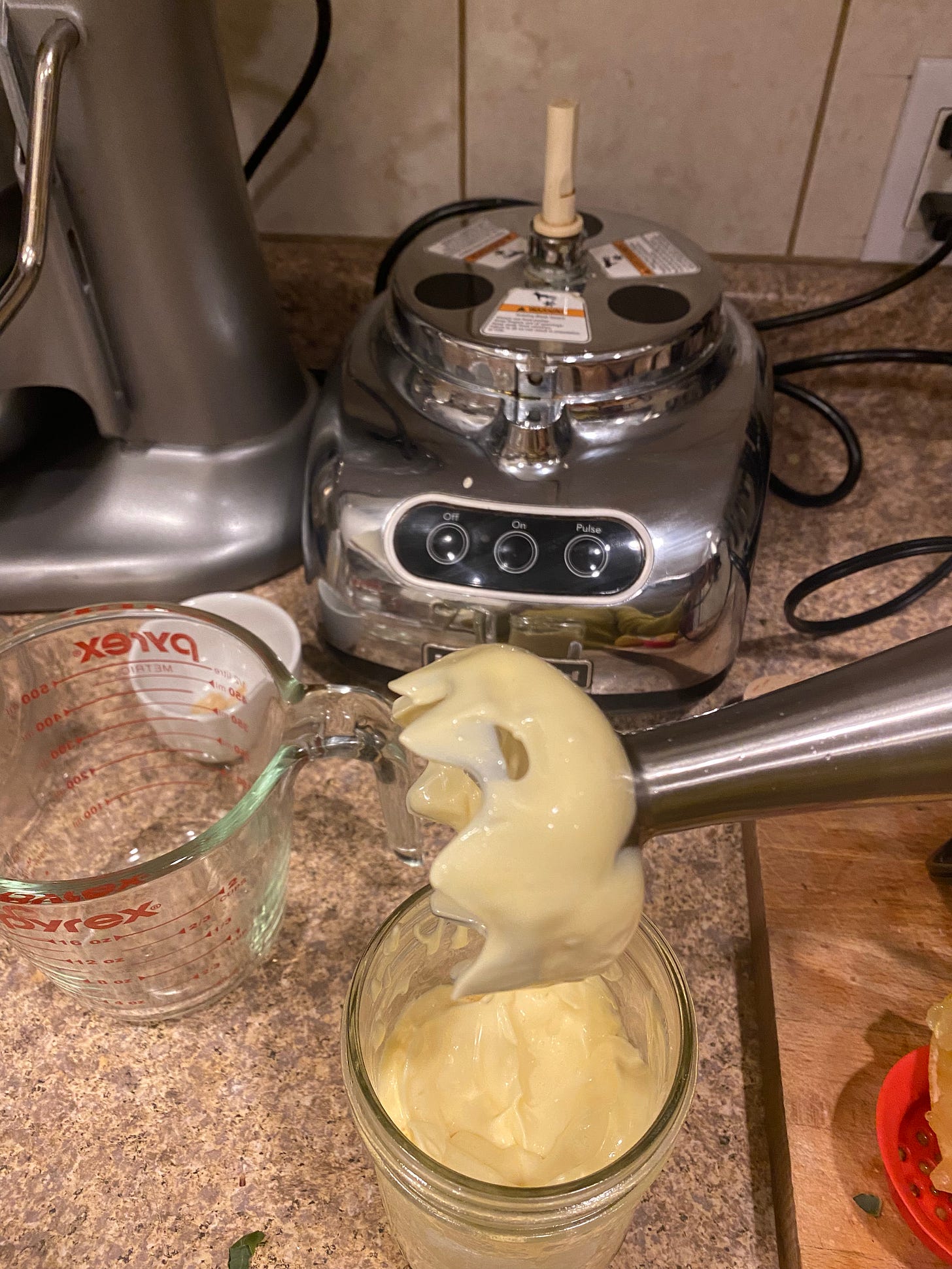 A wide-mouth glass mason jar with mayo in it. Above it, an immersion blender shows the texture of the mayonnaise. The counter behind the jar shows a measuring cup, the base of a food processor, and the edge of a cutting board with a lemon at its corner.