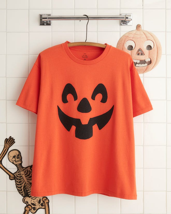 The Pumpkin Tee, a vintage Halloween shirt featuring an iconic jack-o'-lantern pattern and crafted from 100% cotton, hangs on a white wall hook.