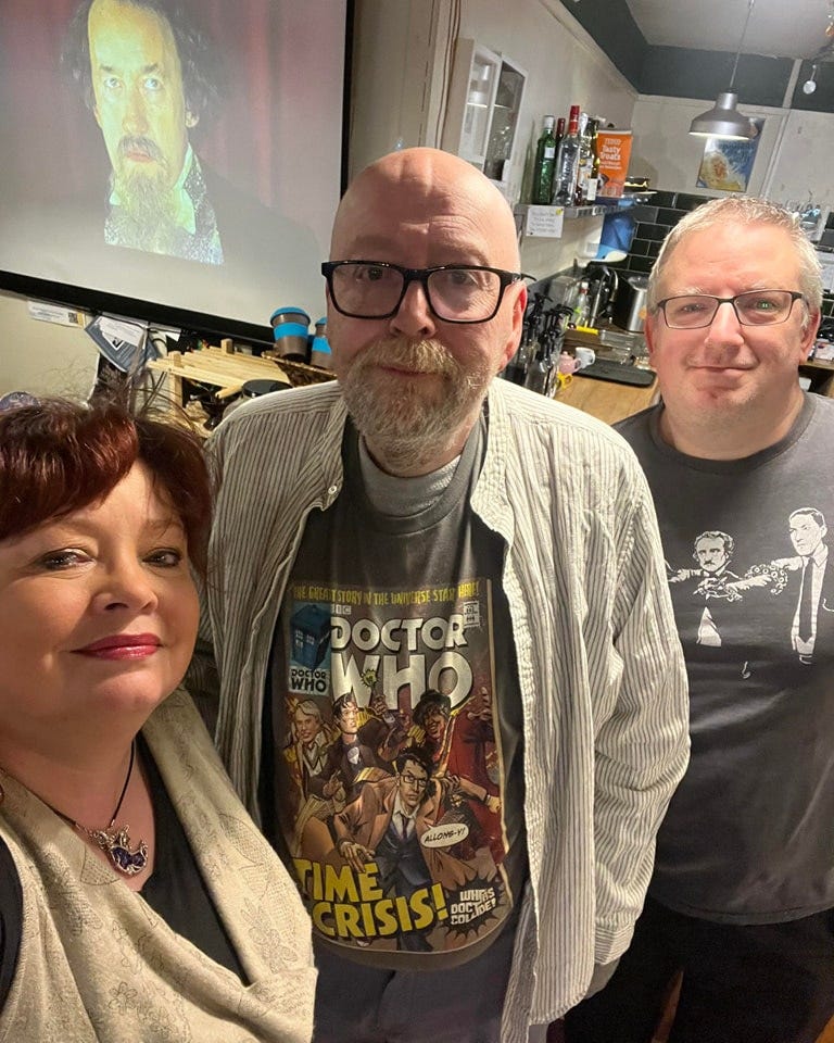Three people, in Script Haven bookshop, left to right Leena Bachelor (former Worcestershire Poet Laureate), Ade Couper (former Worcestershire Poet Laureate) and Damon Lord (current Worcestershire Poet Laureate 2023/24). In the background, a projected image of Simon Callow as Charles Dickens appears.