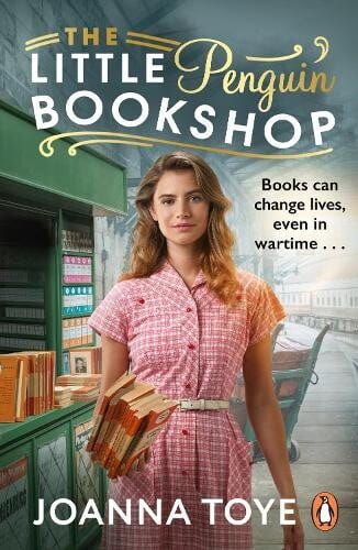 book cover for The Little Penguin Bookshop by Joanna Toye. A woman in a pink gingham dress is holding a stack of books standing in a library