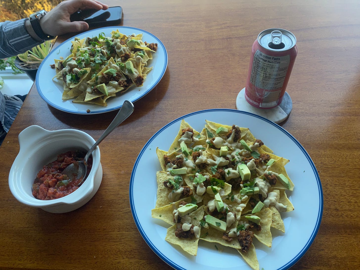Two plates of vegan nachos as described above: lentil meat, queso, avocado, and cilantro on top. There's a white french onion soup bowl between them full of chunky red salsa with a spoon in it. Jeff's hand is just visible at the top left of frame, and a pink can of sparkling water is on the coaster next to the plate in the foreground.