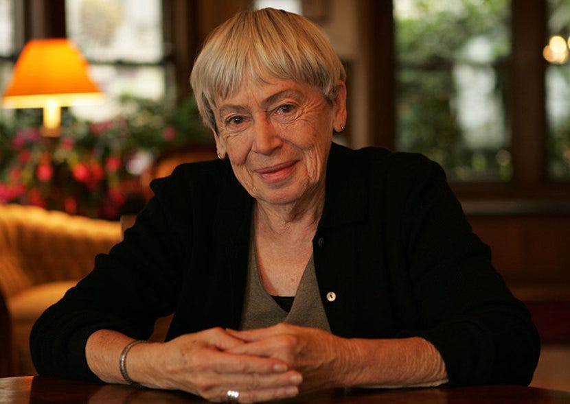 This color photograph shows author Ursula K. Le Guin sitting at a table in her home and smiling at the camera. This color photograph shows author Ursula K. Le Guin sitting at a table in her home and smiling at the camera.