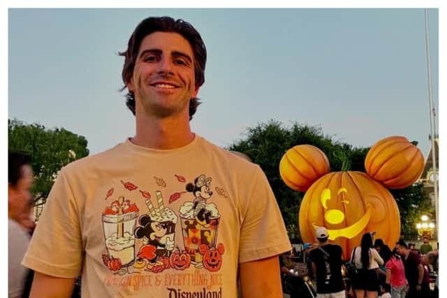 A young, fit man wearing a Disneyland-themed t-shirt stands smiling at Disneyland with a large, illuminated Mickey Mouse pumpkin in the background. The man appears happy and energetic.