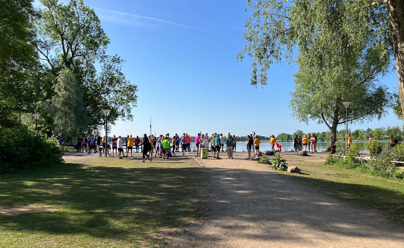 A crowd of parkrunners by a lake, with some shade from tall trees