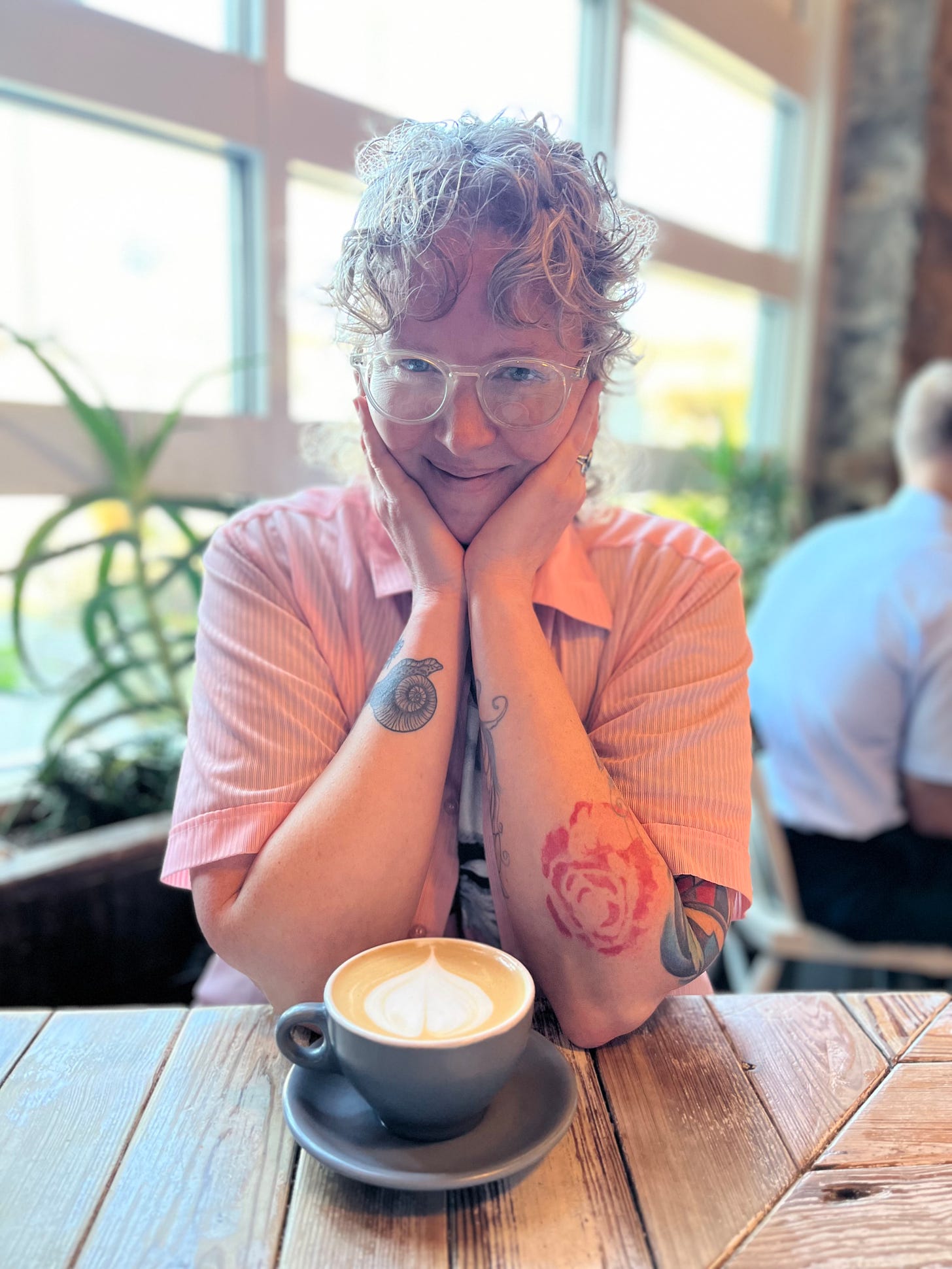 a person with curly hair wearing a light pink button-up gazes at the camera smiling, chin resting in their hands, tattooed arms propped on a wooden table with a latte on it in a sunlit cafe