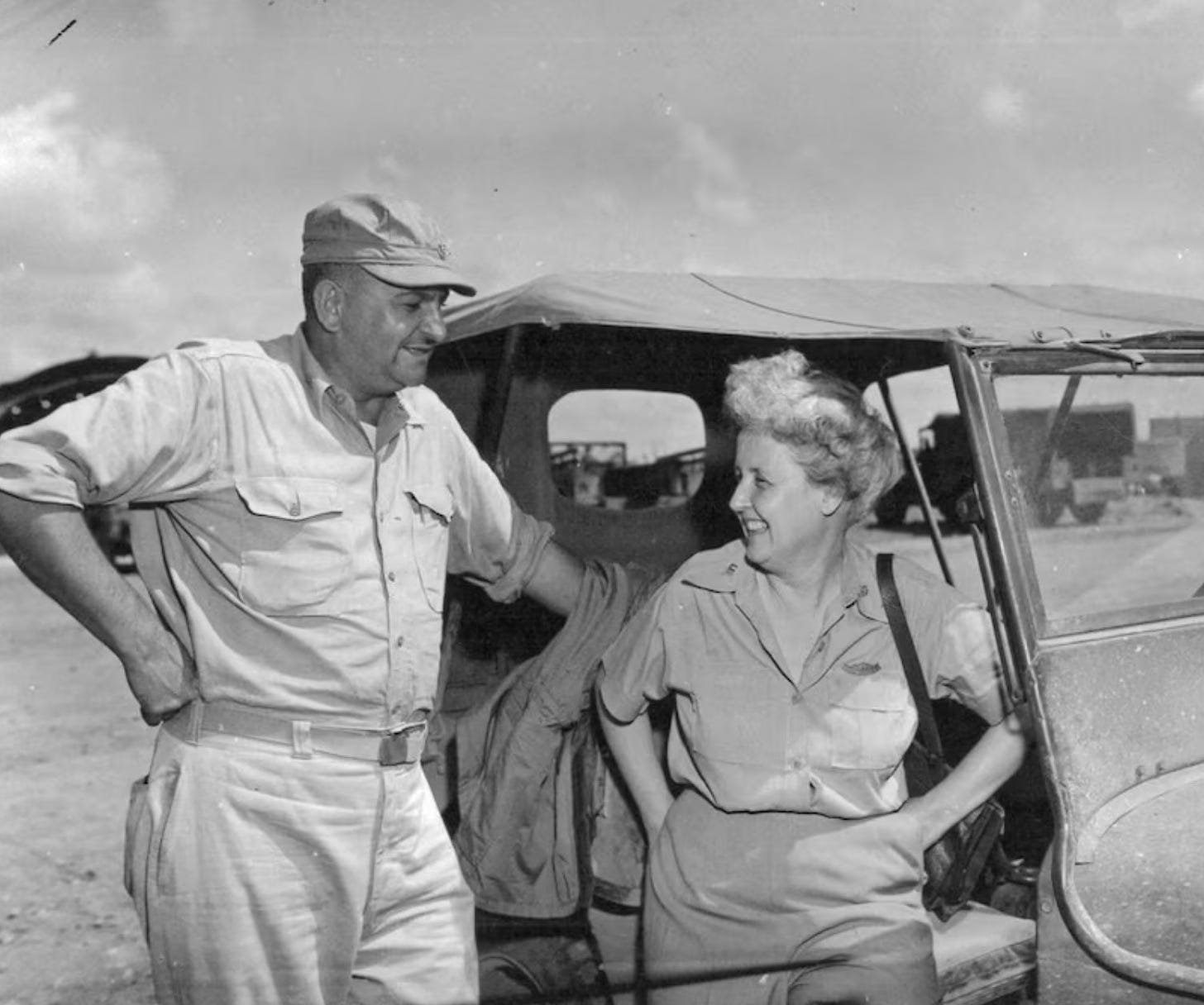 A picture of Barbara with her husband Percy, also a war correspondent.  They are standing next to a jeep, apparently talking and smiling at each other.