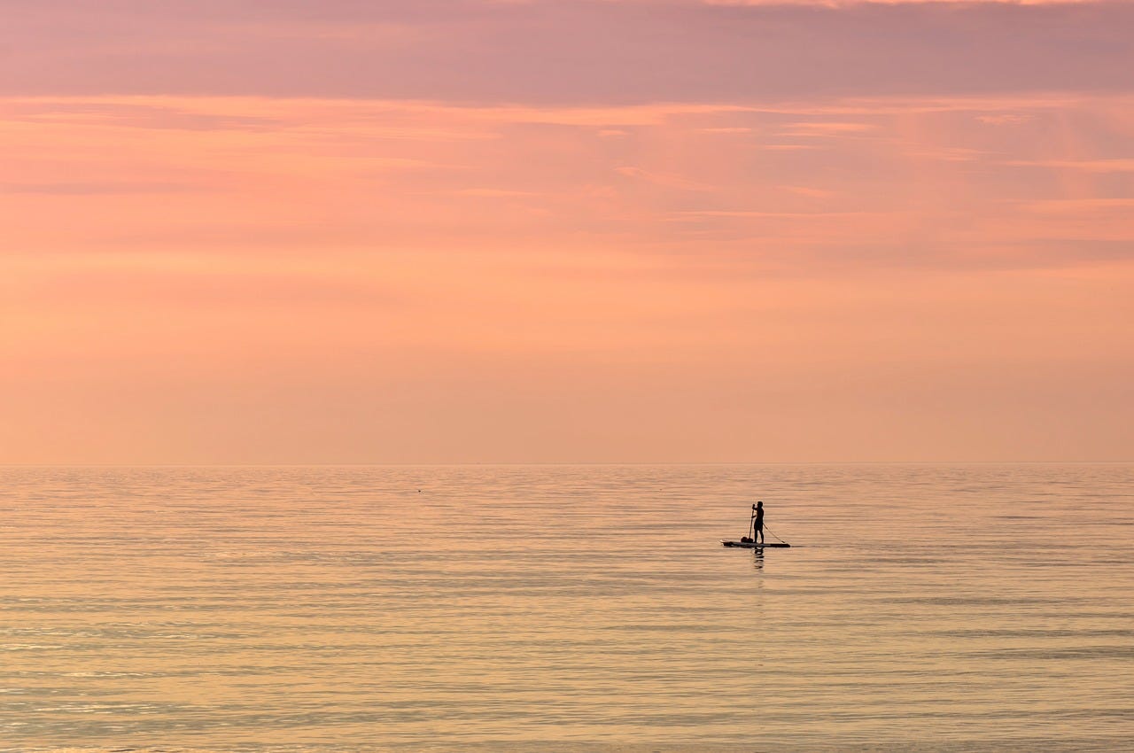 silhouetted man rows a raft in the middle of a still ocean under a pink sky