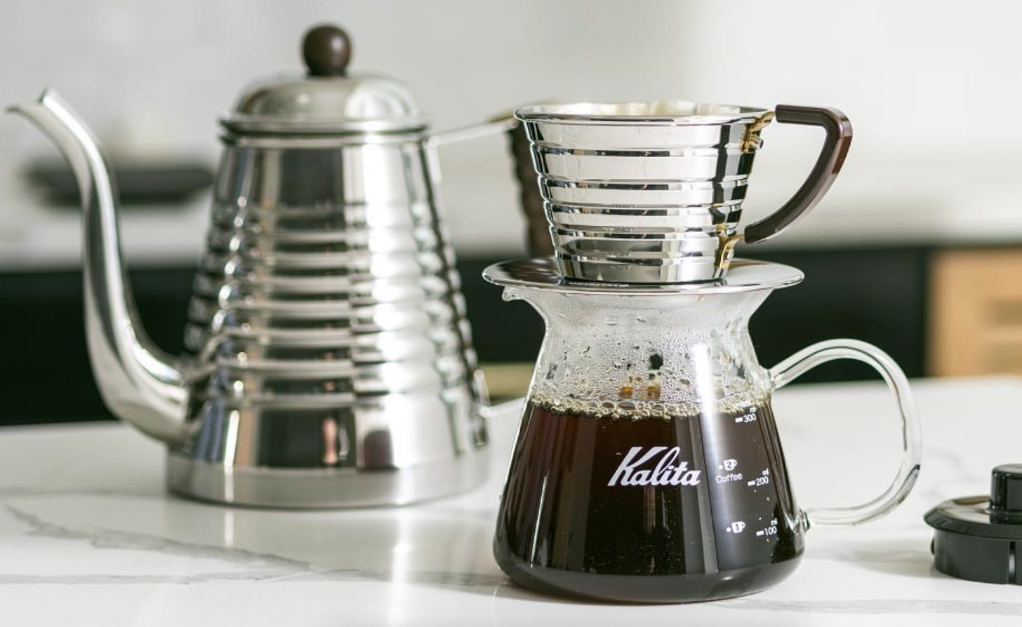 A silver water kettle is blurred in the background (left) on a white marble countertop. A silver cone-shaped pour over coffee brewing device with ridges called the Kalita Wave sits on a glass Kalita branded coffee beaker filled 2/3rds of the way up.