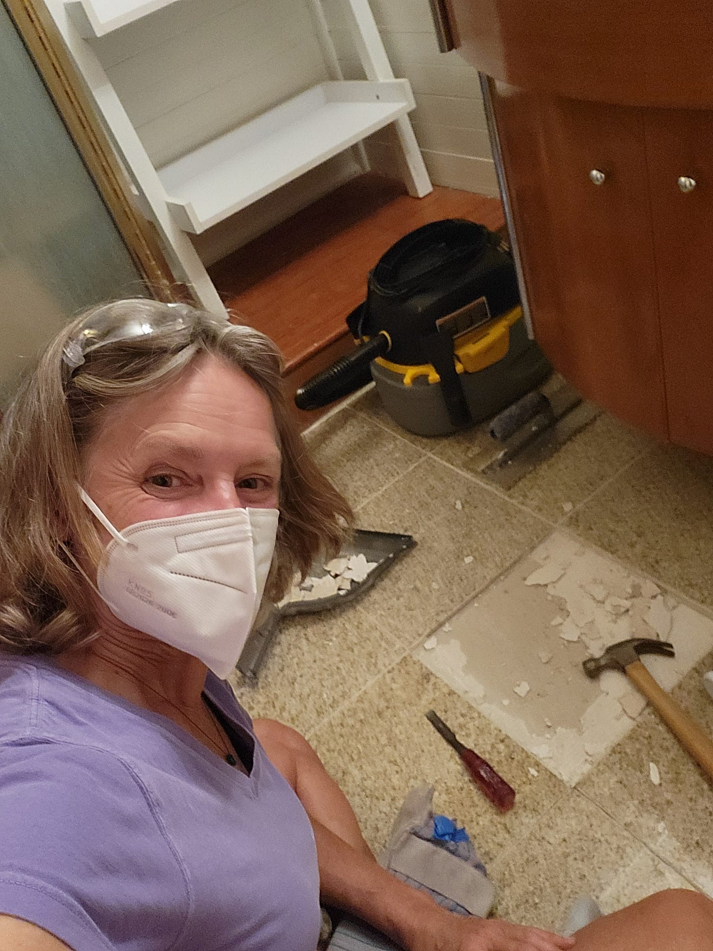 Melanie sitting on a floor replacing a tile. She's smiling behind her mask and protective goggles, and there is a shop vac in the background ready to help clean up!