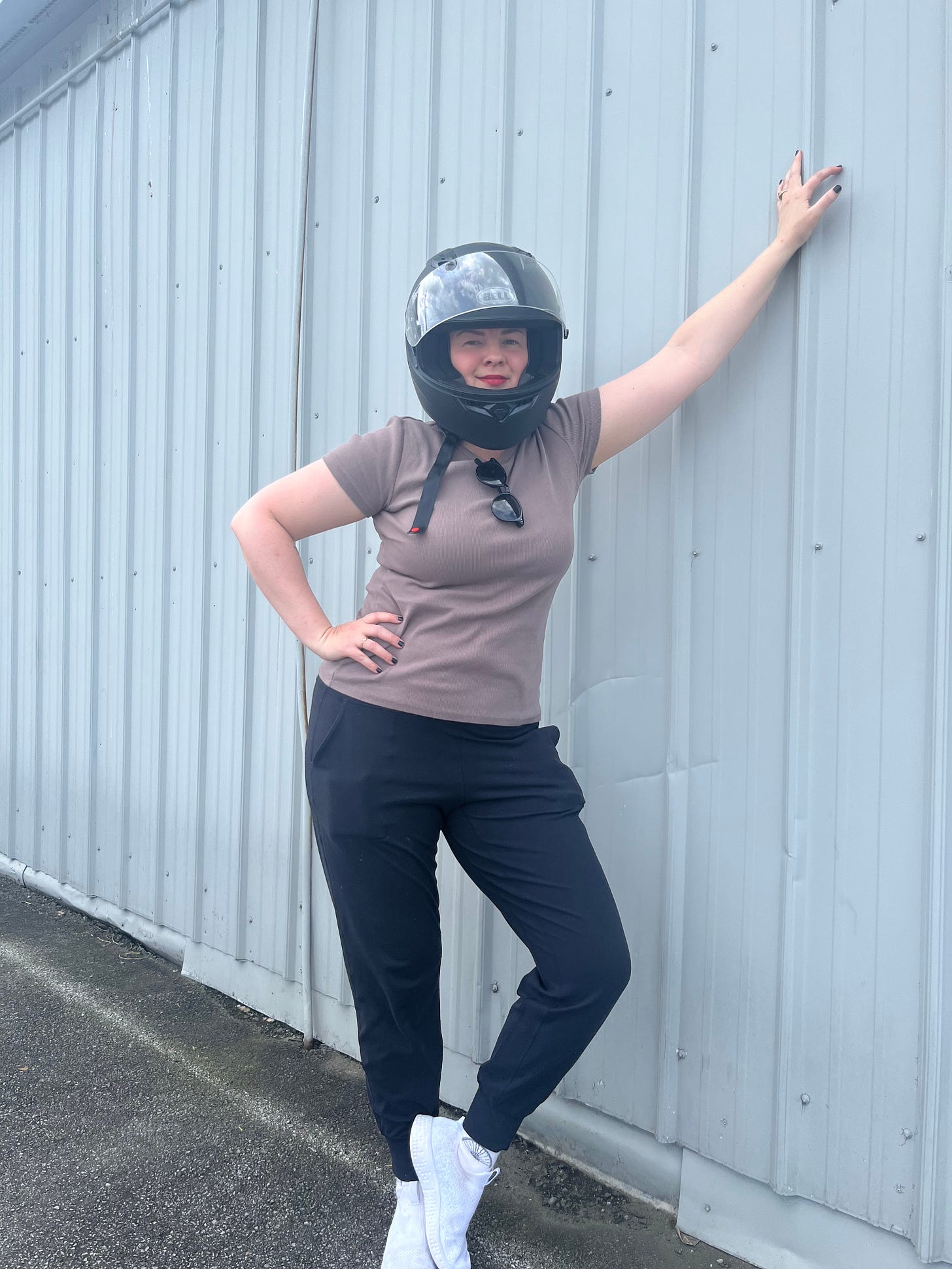 Amber has one hand on her hip and the other propping herself up against a building. She is wearing her motorcycle helmet, looking at the camera, smiling.