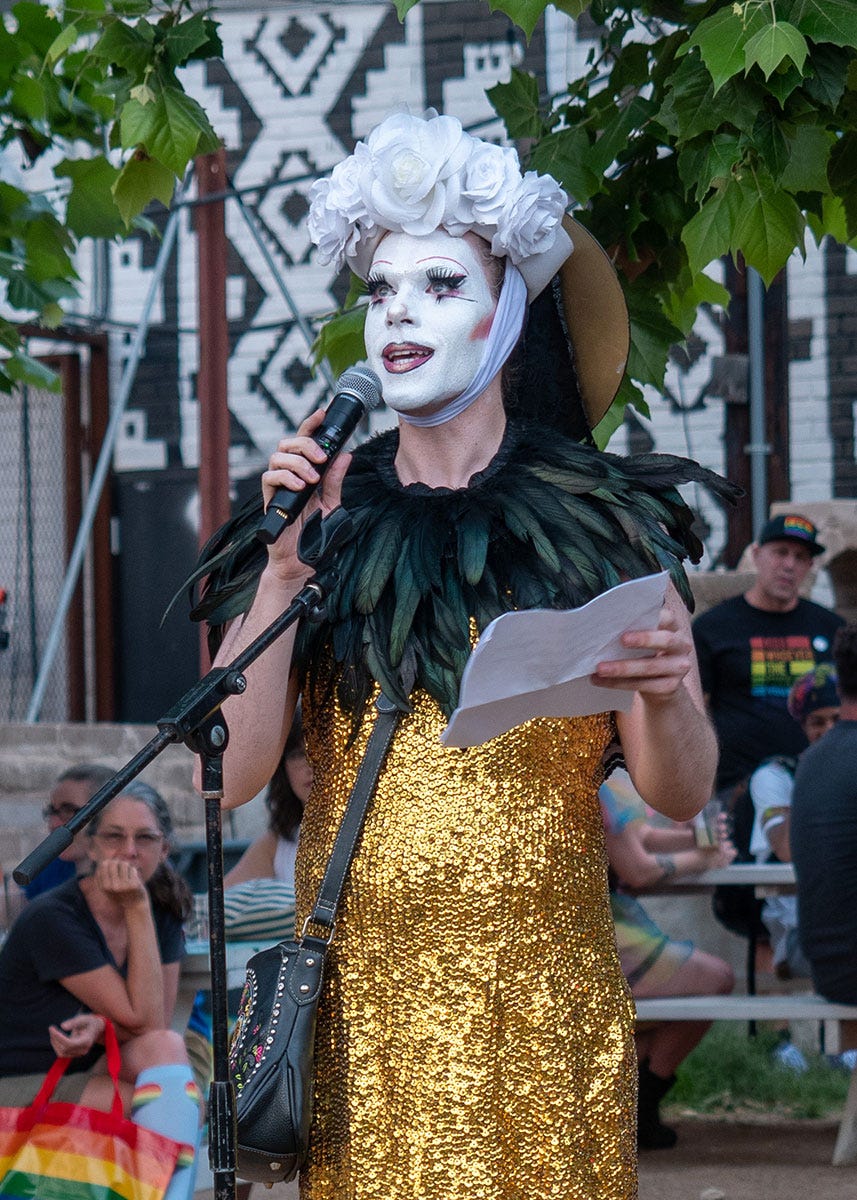 A person gold sequined dress with dark feather color, white face paint, and white rose head piece, speaks into a microphone.