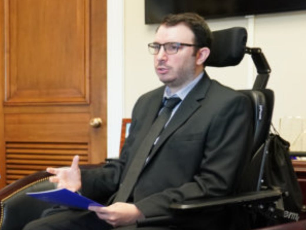 The author, a while male, wearing a black suit with black tie, sitting in a wheelchair. He is holding a blue folder in his left hand. Behind him is a brown wooden door and a white wall.