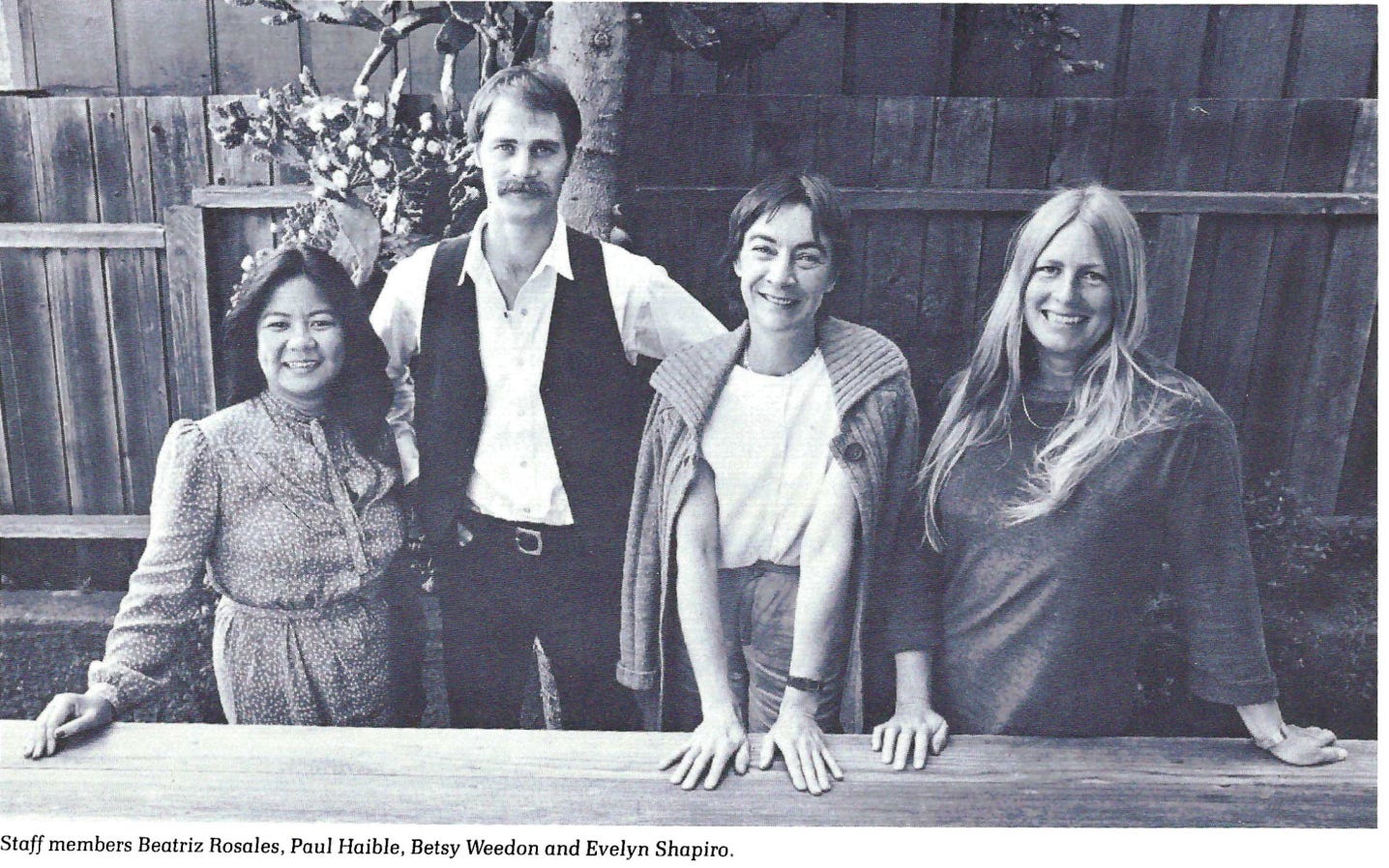 4 people posed together in business casual 70's/80's clothes in front of a tree in a backyard. There is one asian women on the left side and a tall white guy to her right and two white women next to him.