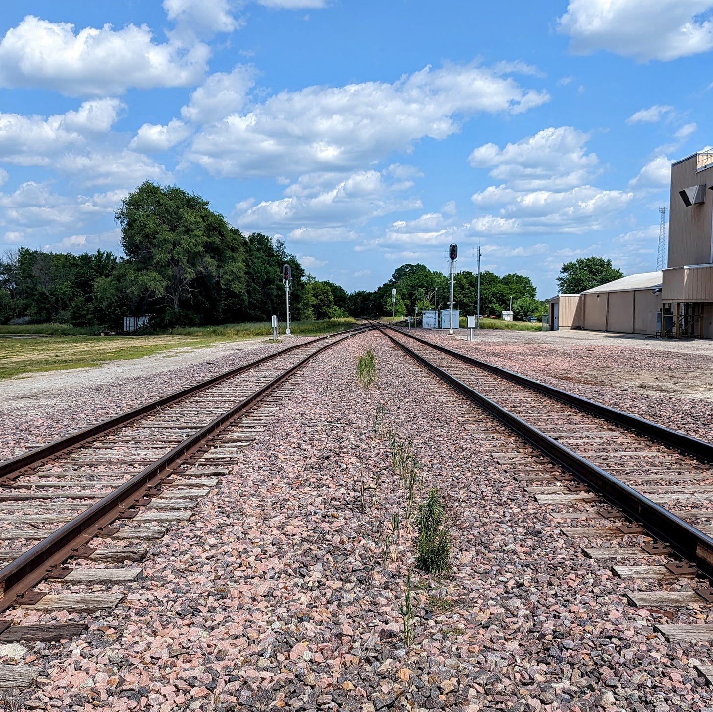 Two rows of train tracks, with gravel between them. Trees are on the left, and a building is on the right.