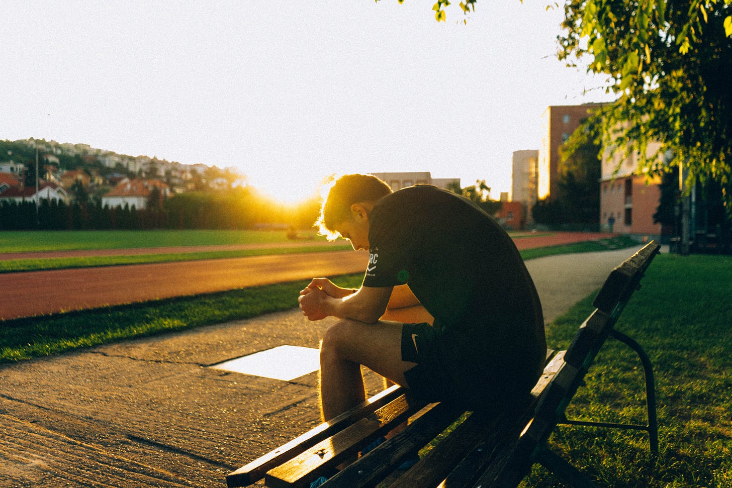 Photo of a person sitting on a bench. They are hunched over, maybe sad or thoughtful. Time of day is sunrise or sunset.