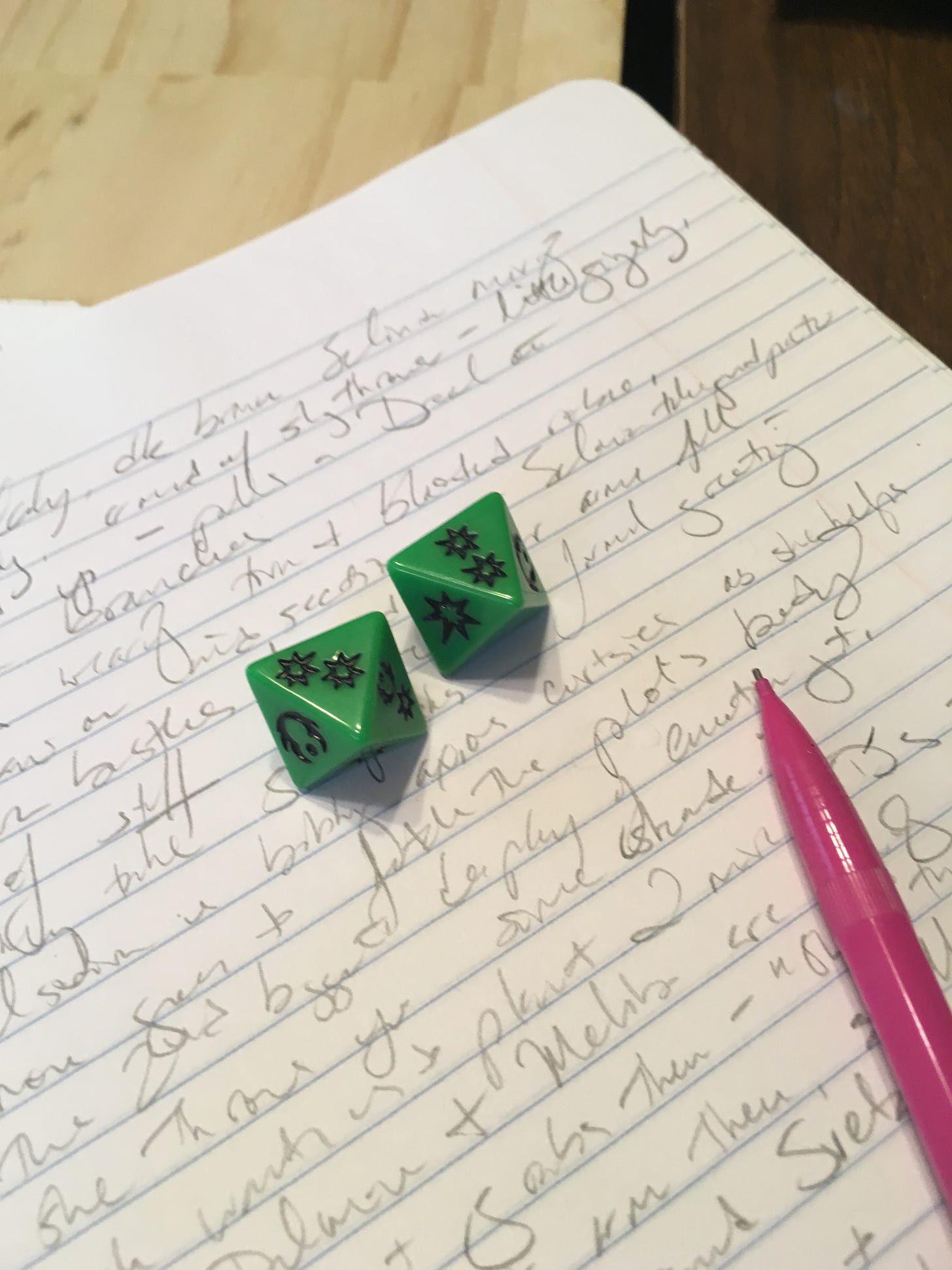 Two green dice and a pink mechanical pencil on top of a much-scribbled-on page of a notebook