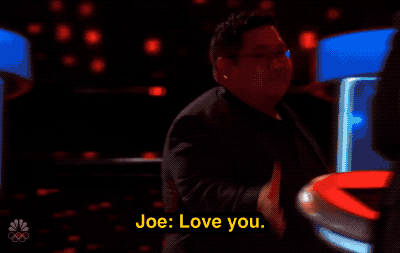 GIF of a dark gameshow set in which Joe Fejeran, a CHamoru man, is walking as a caption in yellow reads "Joe: Love you" while he passes by the tall blonde celebrity lesbian Jane Lynch in a suit as she blows him a kiss.