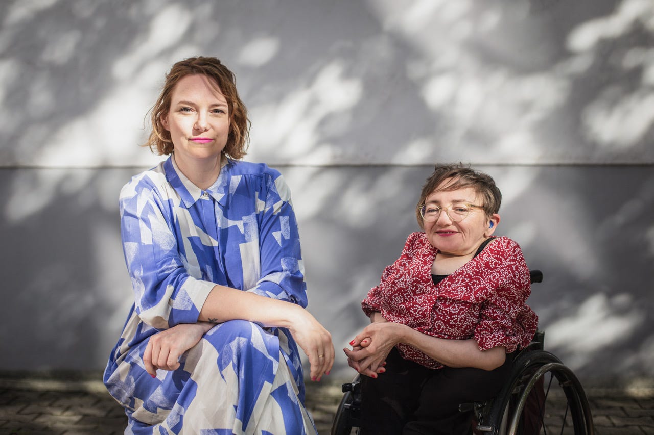 Vor einer einer weißen Hauswand, auf der der Schatten eines Baumes sichtbar ist, sitzen von links nach rechts Mareice Kaiser und Rebecca Maskos. Mareice trägt ein blau-weiß gemustertes Kleid und hat die Hände im Schoß liegen. Rebecca sitzt in einem elektrischen Rollstuhl und trägt eine rote Bluse mit weißem Muster. Sie hat die Hände im Schoß gefaltet. Beide lächeln in die Kamera.