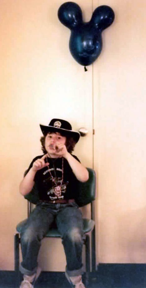 Image description: A ten year old kid sitting on a chair in front of a hotel room door wearing cuffed jeans, a black t-shirt that says "Secondi Bros Truck Stop, which is where their father had worked their whole life at that point, and white Nike sneakers with a blue stripe. They had a necklace with a Native American motif from Frontierland, as well as a black hat that seems to have a badge. They're playing a mouth harp also acquired in Frontierland. There's a dark blue mouse ears balloon tied to the doorknob.