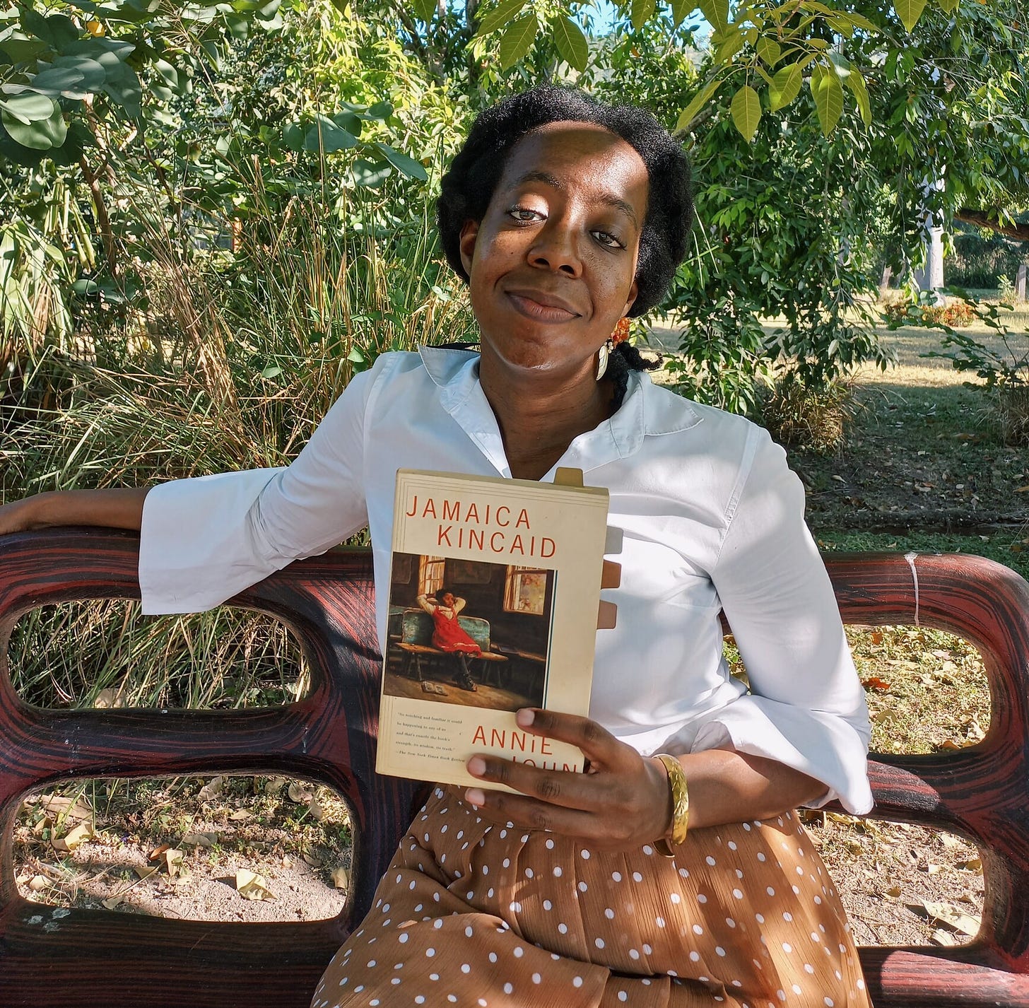 A black woman with natural hair in a loose ponytail, wearing a brown and white polka dotted skirt and long sleeve white shirt holds a paperback copy of Annie John in a red bence under a tree surrounded by greenery. She stares at the camera as sunlight streaks across her face.