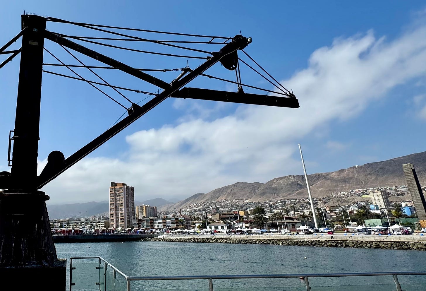 Foto de Antofagasta. En primer plano, maquinaria del puerto histórico, y un poco de mar. Después, ciudad moderna. A lo lejos, barrios humildes en el cerro y la Sierra de la Costa. Foto propia.