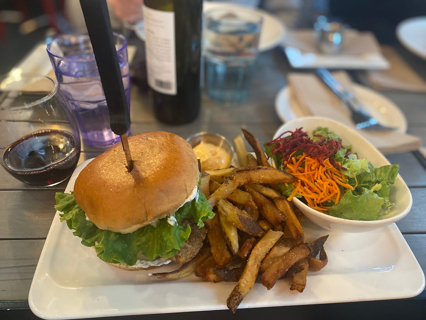 A rectangular plate with a fried oyster mushroom burger, fries, and salad with shredded beet and carrot. A knife is stabbed through the centre of the burger, and there's a glass of wine and a glass of water beside the plate. In the background you can see a wine bottle and a plate across the table.