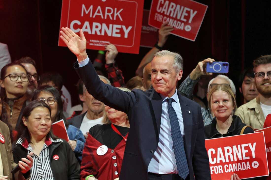 In this photo, Canadian Prime Minister Mark Carney stands outdoors behind a lectern during an election rally in Windsor, Ontario, on April 26, 2025. He's surrounded by people, two of whom hold signs saying "Never 51."
