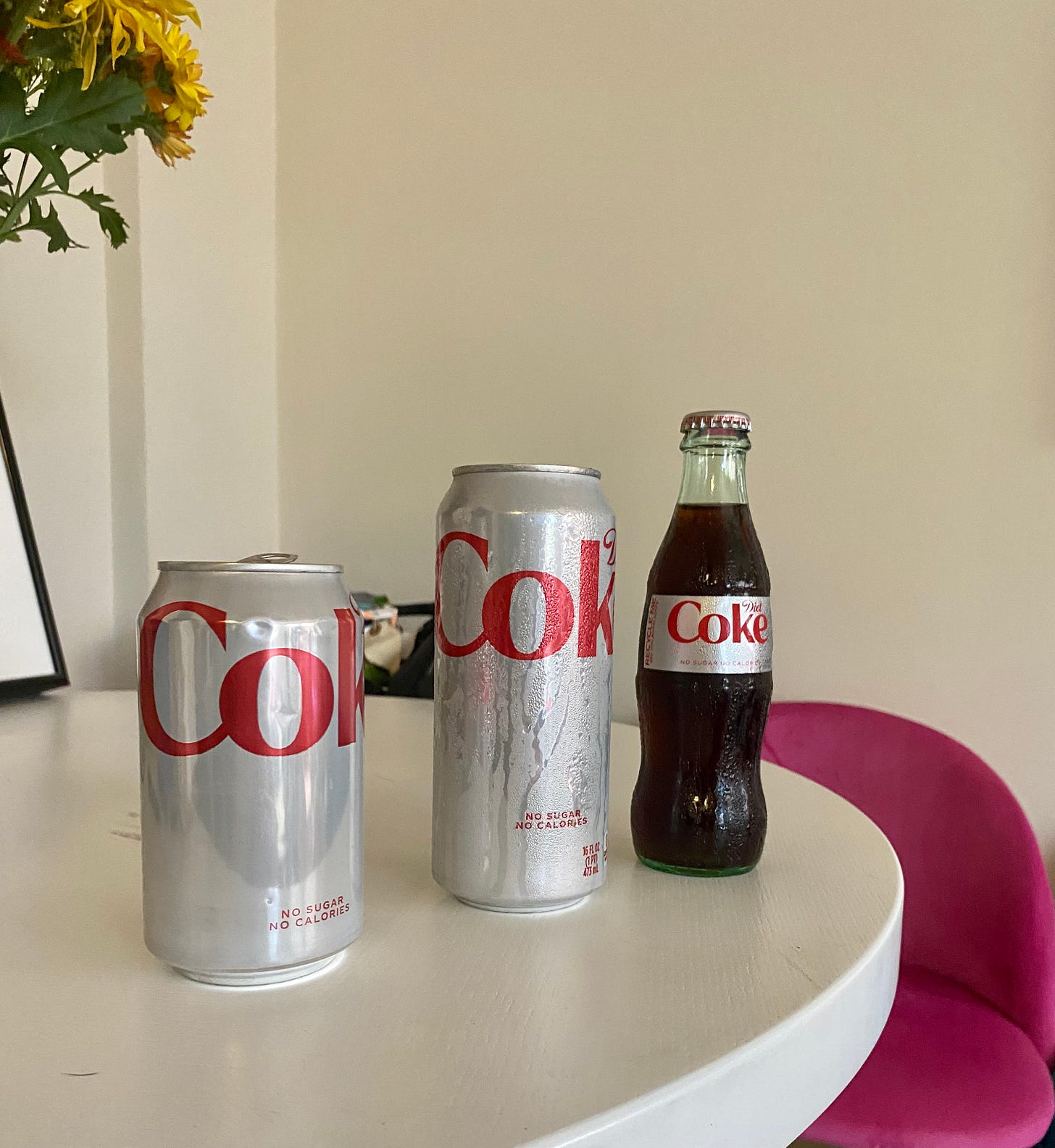 A regular can of Diet Coke, a tall boy can of Diet Coke, and a mini glass bottle Diet Coke on a table