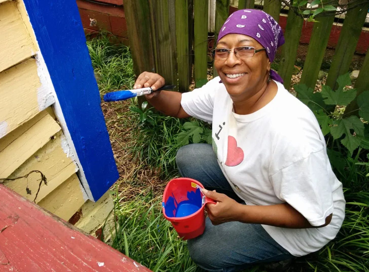 A woman crouching to paint the bright blue trim of her yellow house smiles broadly.