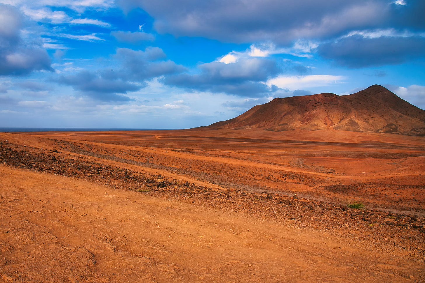 A wide, arid red landscape with volcanic rock and rust-colored soil stretches across Sal Island, Cape Verde, beneath a dramatic blue sky scattered with clouds. A low mountain rises to the right, casting soft shadows across the terrain. A wide, arid red landscape with volcanic rock and rust-colored soil stretches across Sal Island, Cape Verde, beneath a dramatic blue sky scattered with clouds. A low mountain rises to the right, casting soft shadows across the terrain.