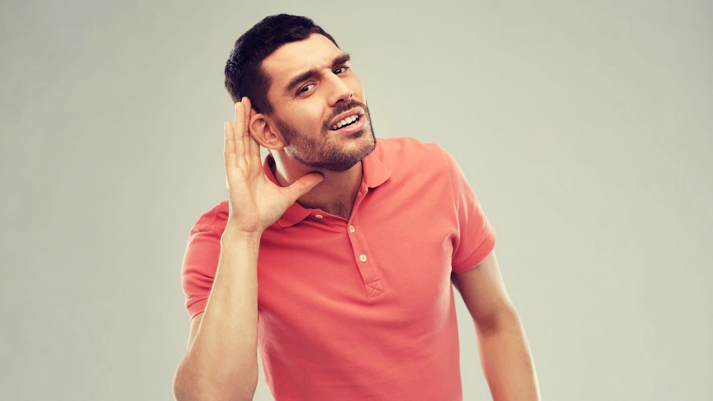 A man in a coral polo shirt leaning forward with one hand cupped to his ear, as if trying to listen.